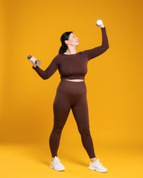 Plus size woman in gym clothes with dumbbells on orange background