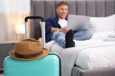 Suitcase with hat and man using laptop on bed in his hotel room, selective focus