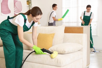 Professional janitor steam cleaning sofa in room