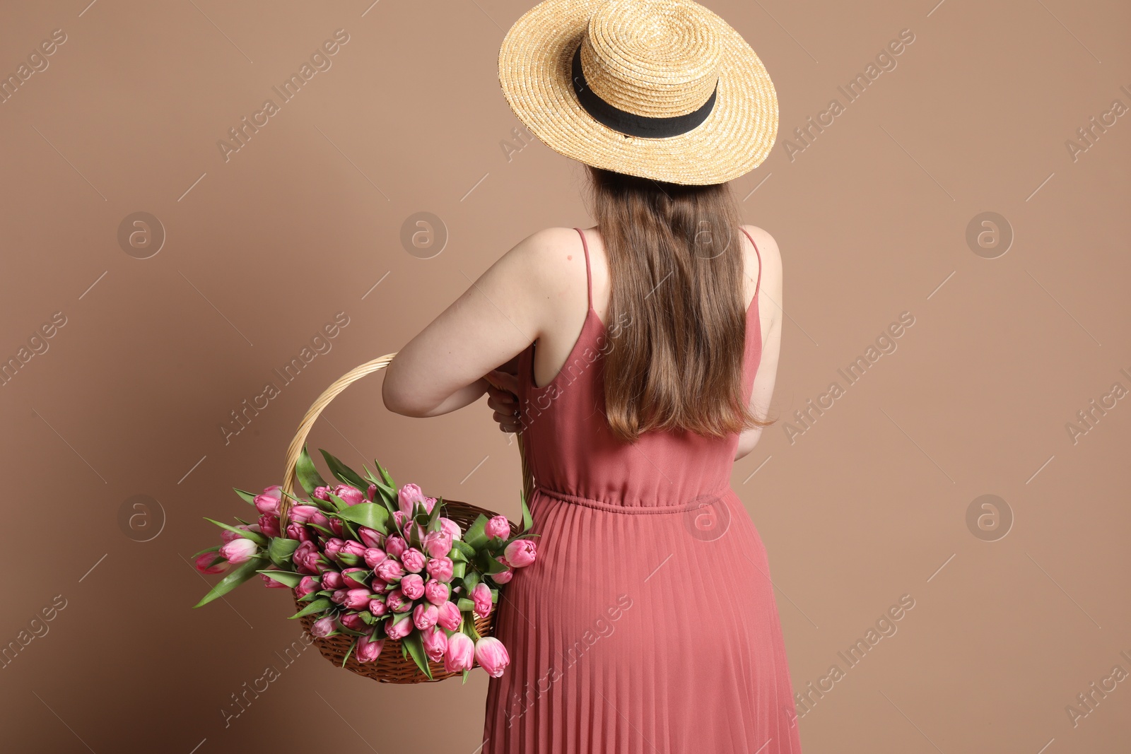 Photo of Woman holding wicker basket with bouquet of beautiful tulips on dark beige background, back view