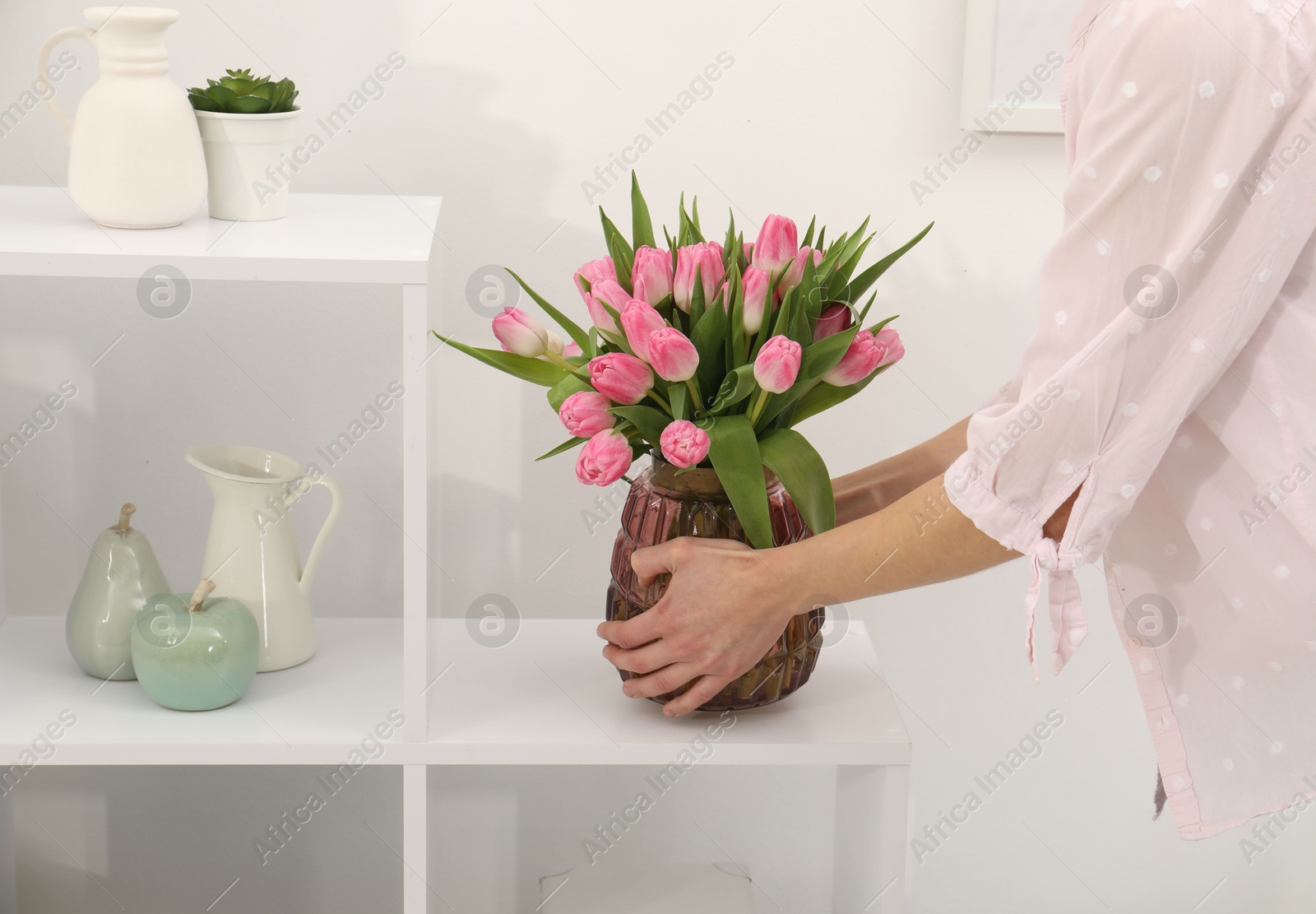 Photo of Woman arranging vase with bouquet of beautiful tulips onto shelving unit indoors, closeup