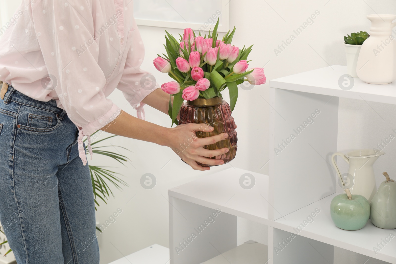 Photo of Woman arranging vase with bouquet of beautiful tulips onto shelving unit indoors, closeup