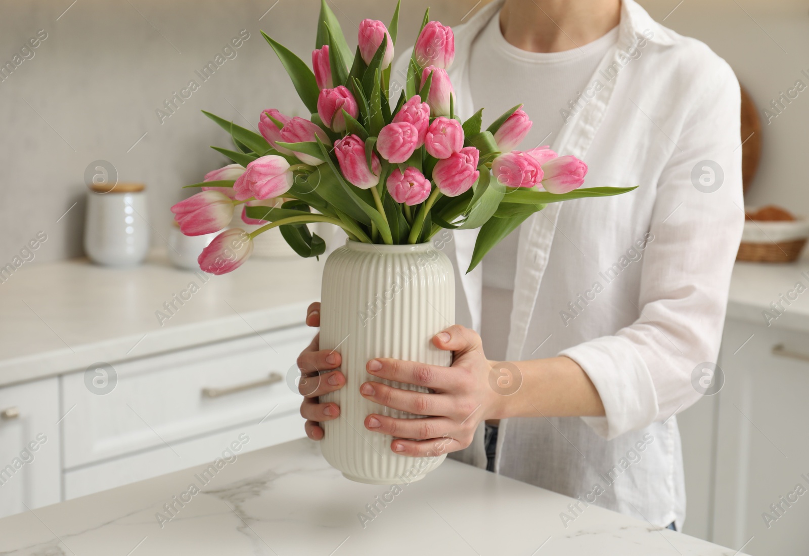 Photo of Woman holding vase with bouquet of beautiful tulips at white marble table in kitchen, closeup