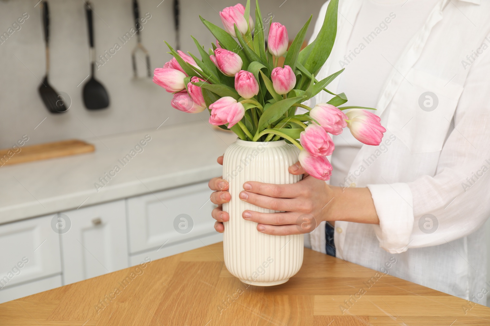 Photo of Woman holding vase with bouquet of beautiful tulips at wooden table in kitchen, closeup