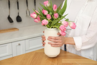 Photo of Woman holding vase with bouquet of beautiful tulips at wooden table in kitchen, closeup