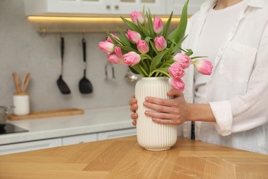 Photo of Woman holding vase with bouquet of beautiful tulips at wooden table in kitchen, closeup. Space for text