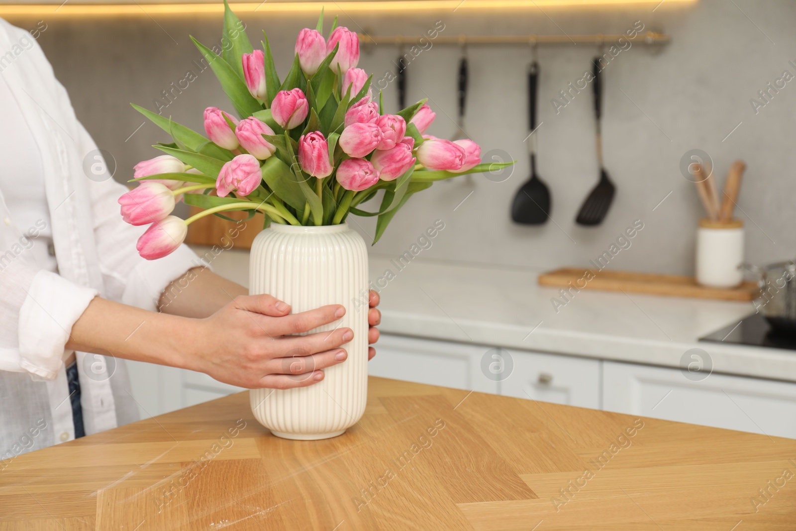 Photo of Woman holding vase with bouquet of beautiful tulips at wooden table in kitchen, closeup. Space for text