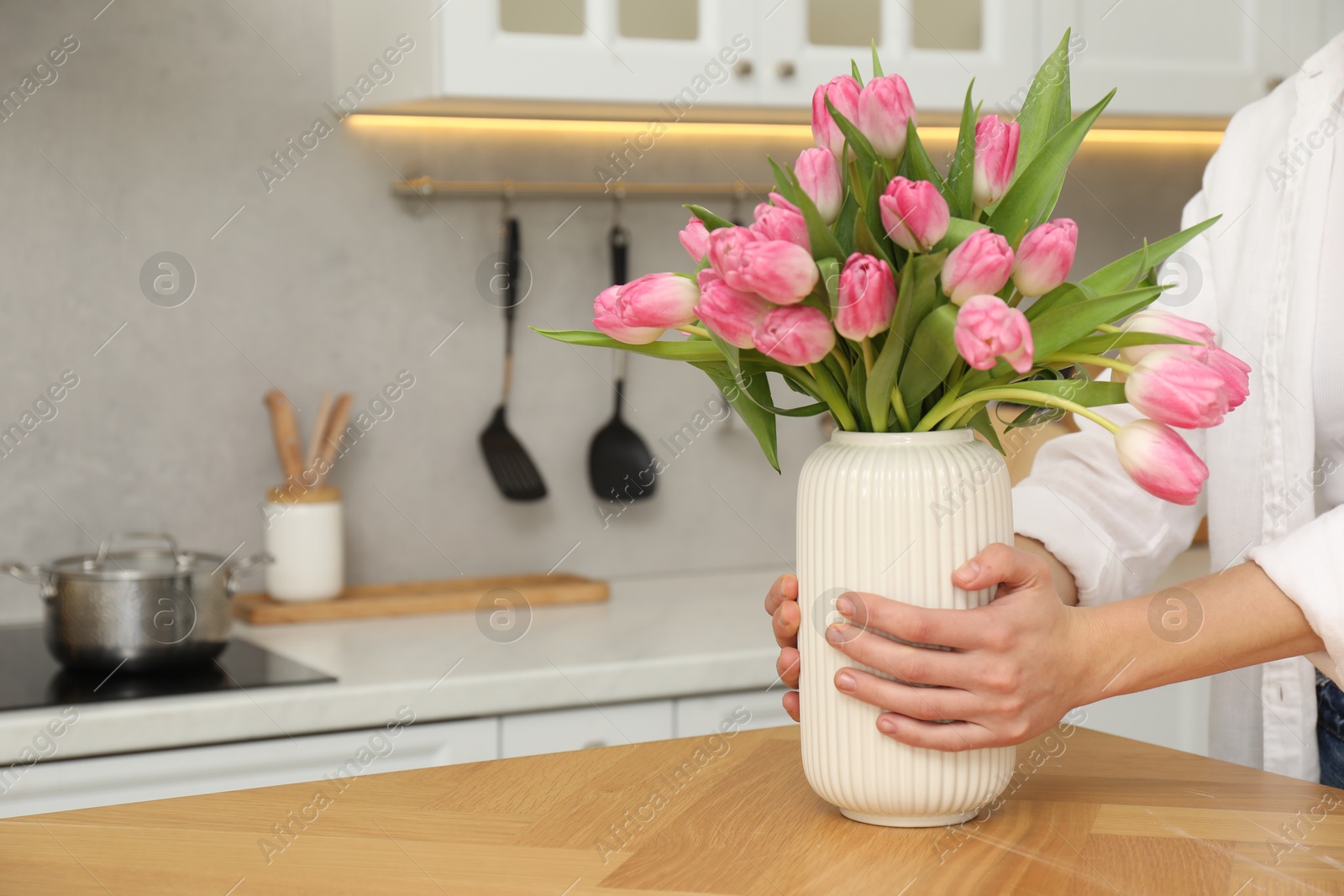 Photo of Woman holding vase with bouquet of beautiful tulips at wooden table in kitchen, closeup. Space for text