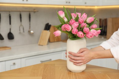 Photo of Woman holding vase with bouquet of beautiful tulips at wooden table in kitchen, closeup. Space for text