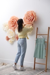 Woman decorating wall with beautiful paper flowers at home, back view