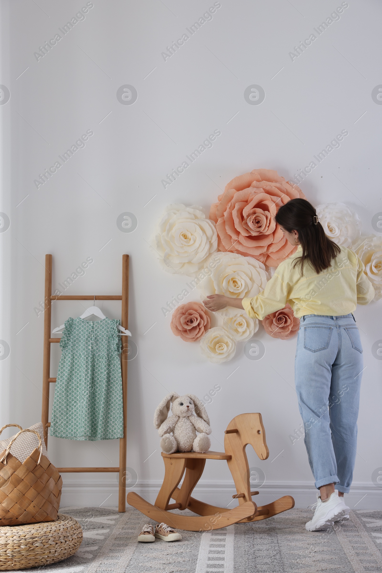 Photo of Woman decorating wall with beautiful paper flowers in child's room, back view