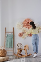 Woman decorating wall with beautiful paper flowers in child's room, back view