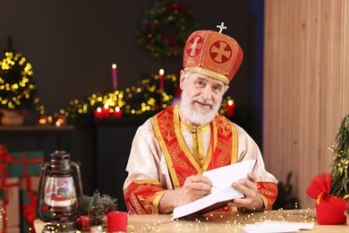 Saint Nicholas with notebook at desk in room decorated for Christmas