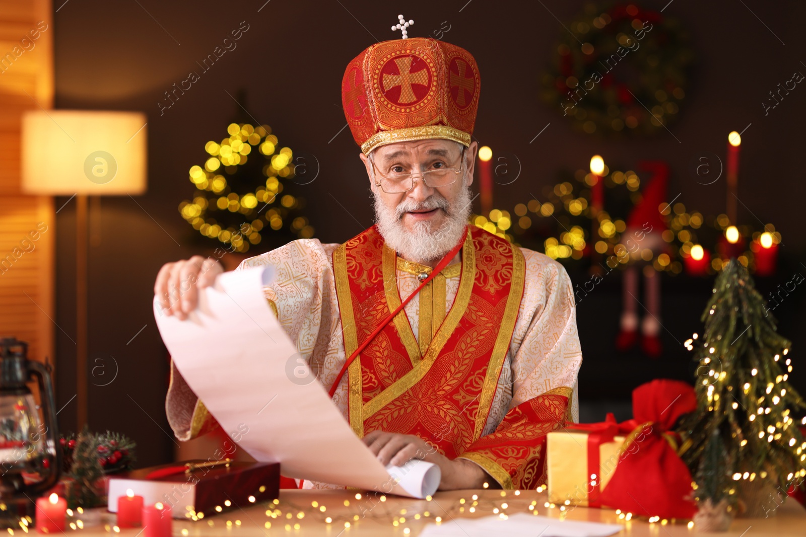 Photo of Saint Nicholas with gift list at desk in room decorated for Christmas