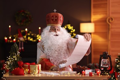 Saint Nicholas with gift list at desk in room decorated for Christmas