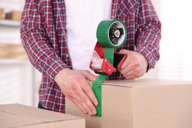 Photo of Man using dispenser with adhesive tape to seal cardboard box indoors, closeup