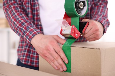 Photo of Man using dispenser with adhesive tape to seal cardboard box indoors, closeup
