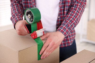 Photo of Man using dispenser with adhesive tape to seal cardboard box indoors, closeup
