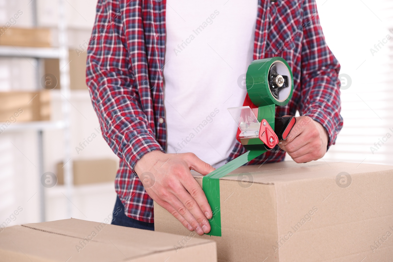 Man using dispenser with adhesive tape to seal cardboard box indoors, closeup Photo of Man using dispenser with adhesive tape to seal cardboard box indoors, closeup