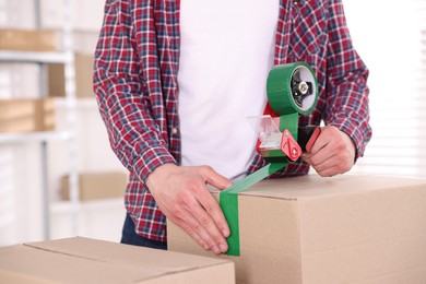 Photo of Man using dispenser with adhesive tape to seal cardboard box indoors, closeup
