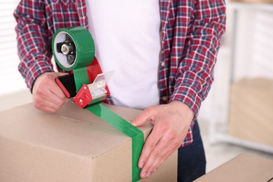 Photo of Man using dispenser with adhesive tape to seal cardboard box indoors, closeup