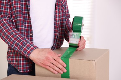Photo of Man using dispenser with adhesive tape to seal cardboard box indoors, closeup