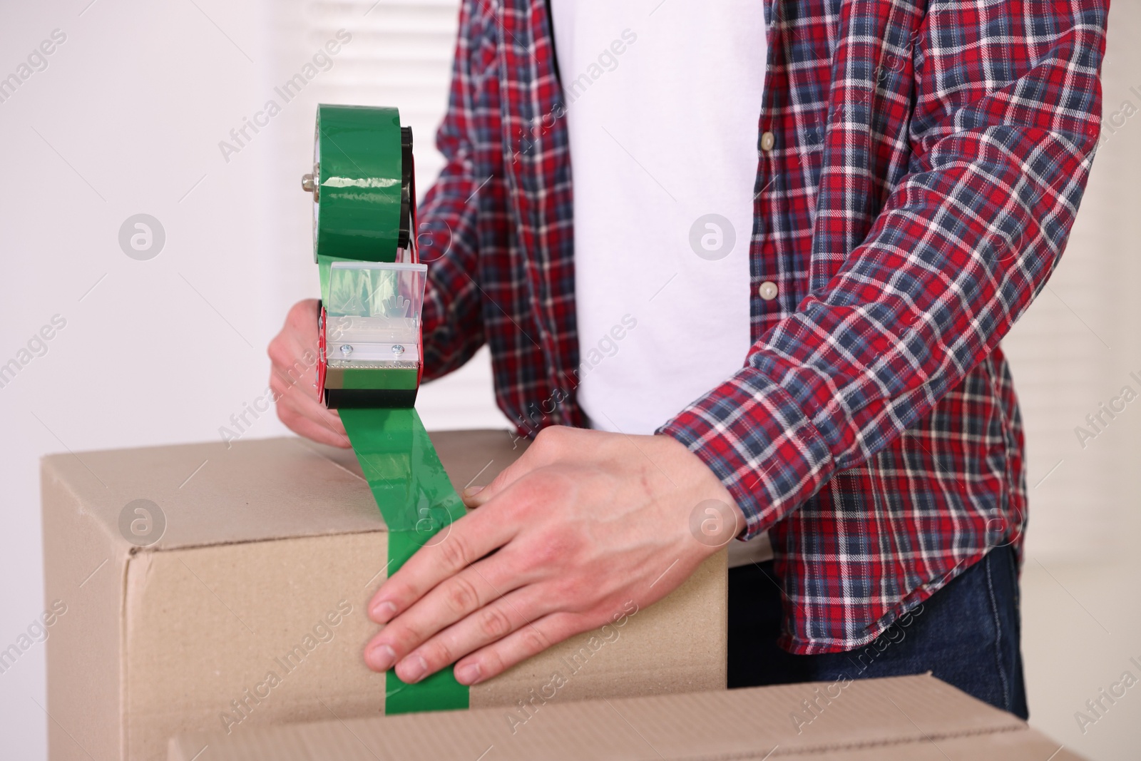 Man using dispenser with adhesive tape to seal cardboard box indoors, closeup Photo of Man using dispenser with adhesive tape to seal cardboard box indoors, closeup