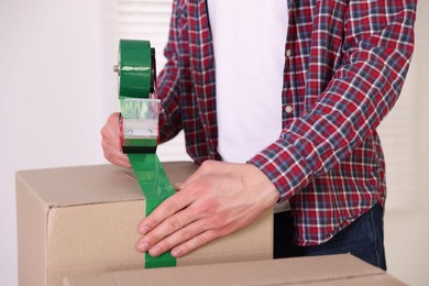 Photo of Man using dispenser with adhesive tape to seal cardboard box indoors, closeup