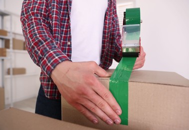 Photo of Man using dispenser with adhesive tape to seal cardboard box indoors, closeup