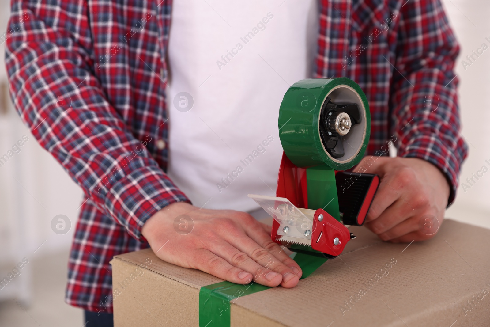 Man using dispenser with adhesive tape to seal cardboard box indoors, closeup Photo of Man using dispenser with adhesive tape to seal cardboard box indoors, closeup