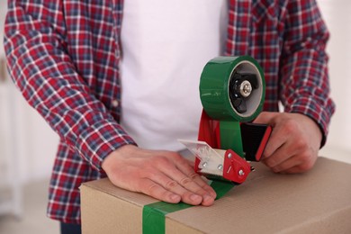 Photo of Man using dispenser with adhesive tape to seal cardboard box indoors, closeup