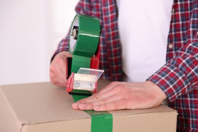 Photo of Man using dispenser with adhesive tape to seal cardboard box indoors, closeup