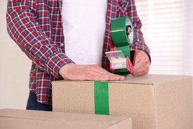Photo of Man using dispenser with adhesive tape to seal cardboard box indoors, closeup