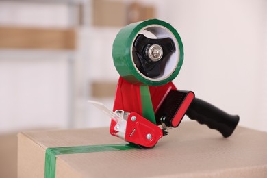 Photo of Dispenser with adhesive tape on cardboard box indoors, closeup