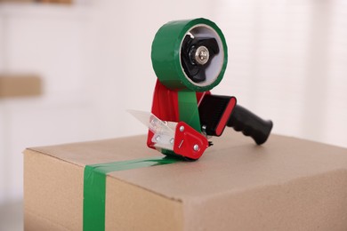 Photo of Dispenser with adhesive tape on cardboard box indoors, closeup