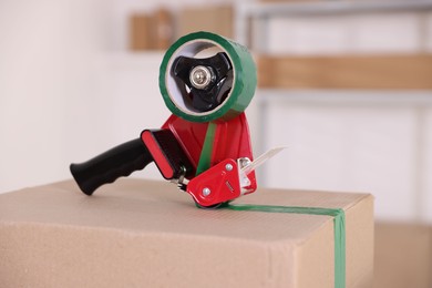 Photo of Dispenser with adhesive tape on cardboard box indoors, closeup