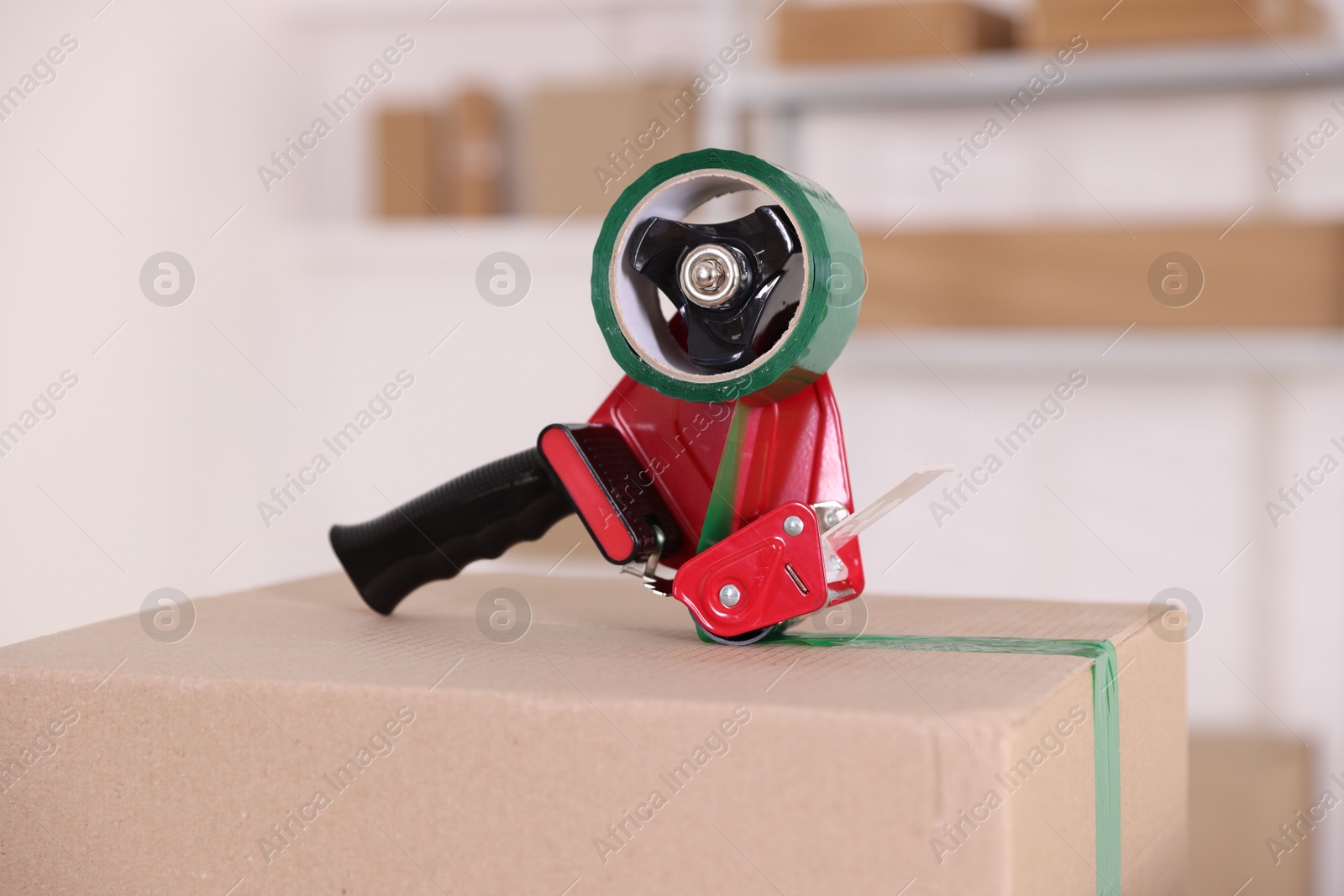 Photo of Dispenser with adhesive tape on cardboard box indoors, closeup