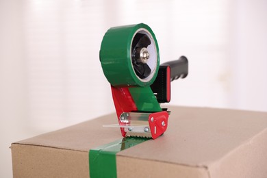 Photo of Dispenser with adhesive tape on cardboard box indoors, closeup