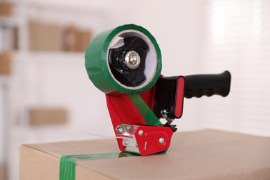 Photo of Dispenser with adhesive tape on cardboard box indoors, closeup