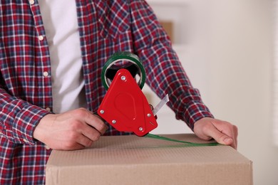 Photo of Man using dispenser with adhesive tape to seal cardboard box indoors, closeup
