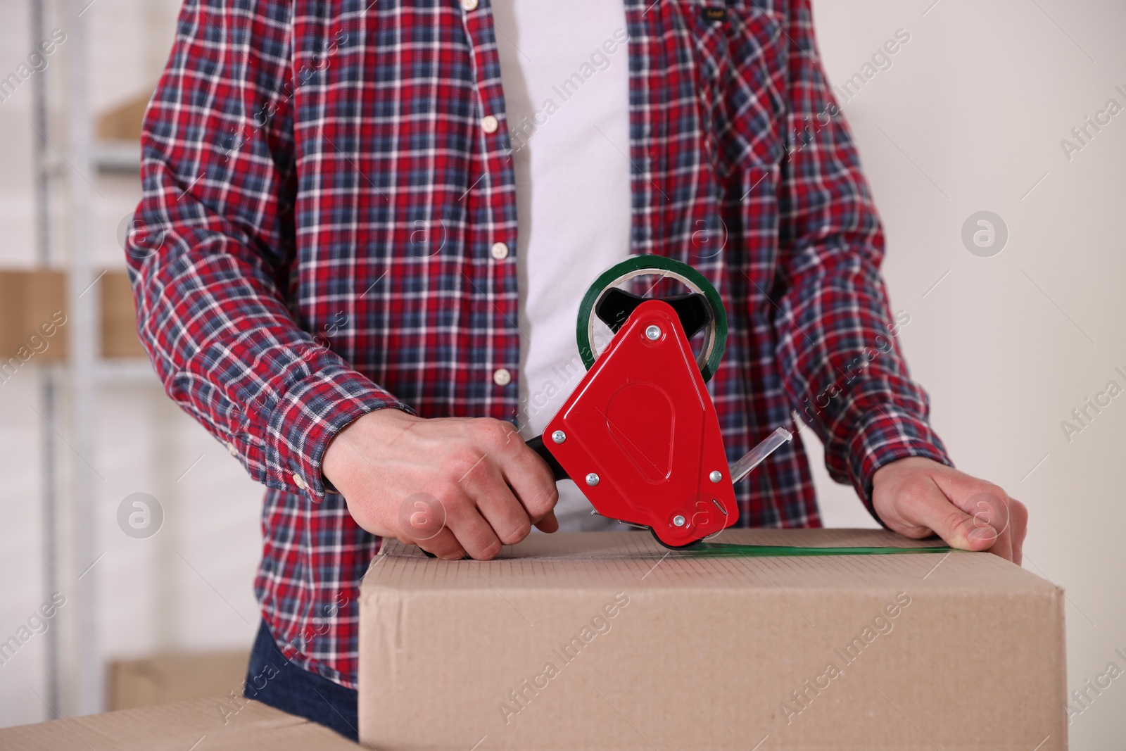 Man using dispenser with adhesive tape to seal cardboard box indoors, closeup Photo of Man using dispenser with adhesive tape to seal cardboard box indoors, closeup