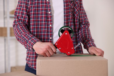 Photo of Man using dispenser with adhesive tape to seal cardboard box indoors, closeup