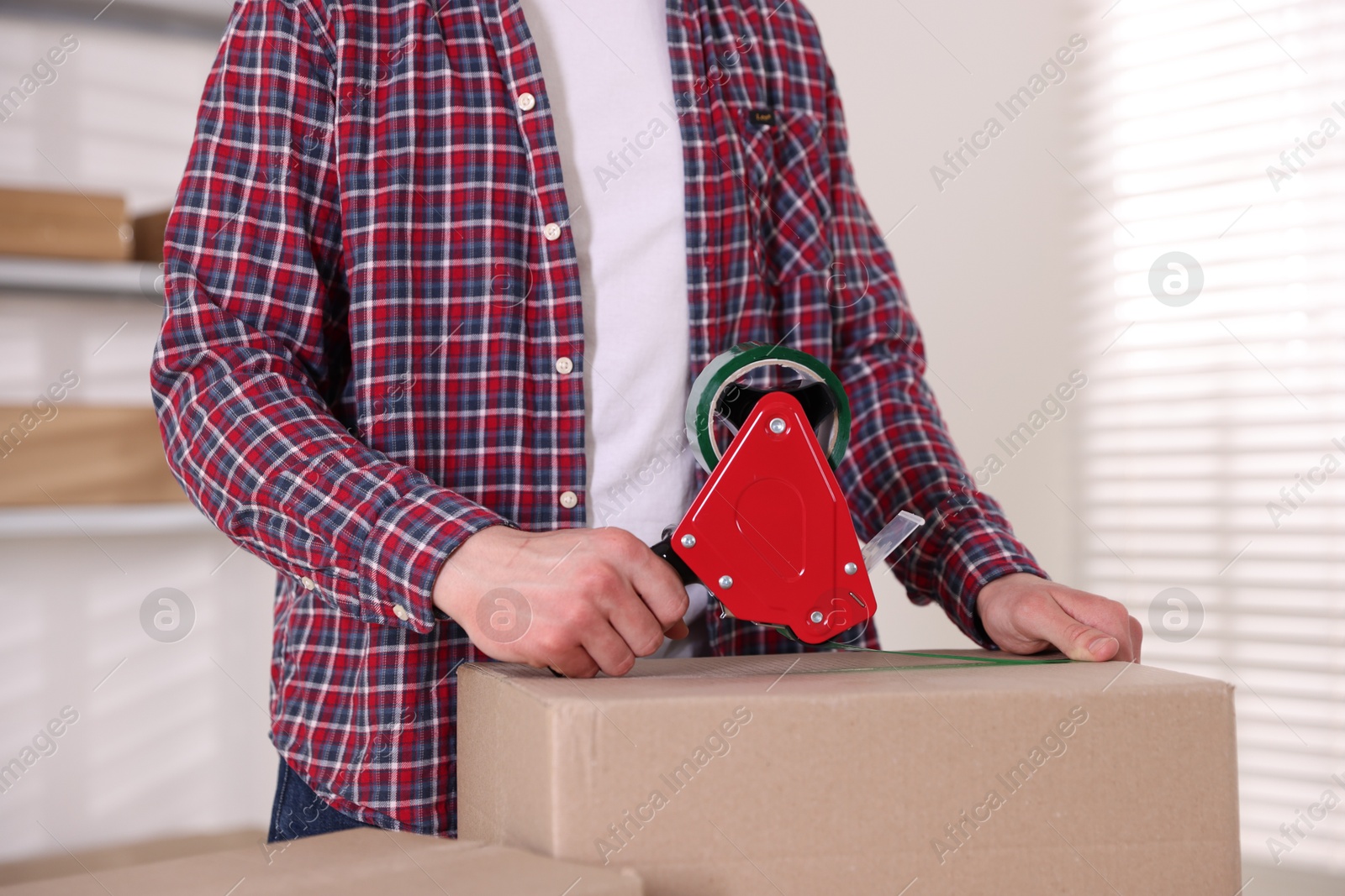 Man using dispenser with adhesive tape to seal cardboard box indoors, closeup Photo of Man using dispenser with adhesive tape to seal cardboard box indoors, closeup