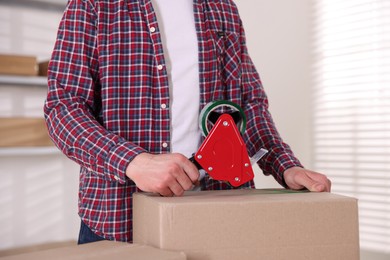 Photo of Man using dispenser with adhesive tape to seal cardboard box indoors, closeup