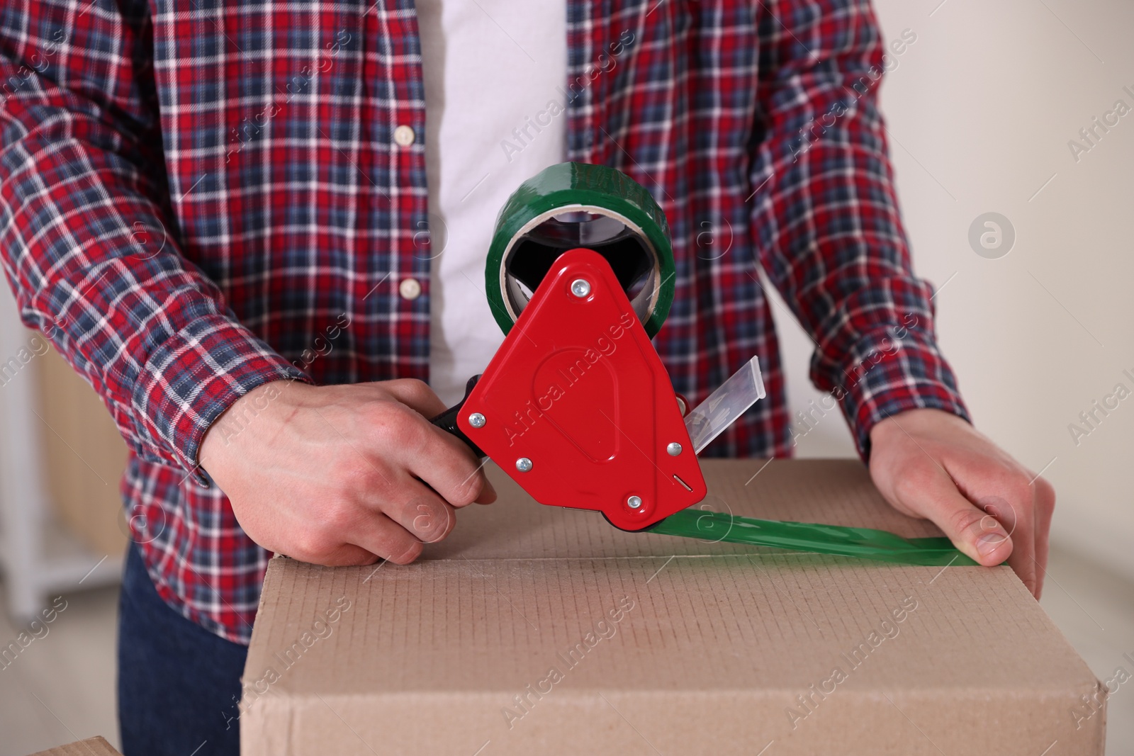 Man using dispenser with adhesive tape to seal cardboard box indoors, closeup Photo of Man using dispenser with adhesive tape to seal cardboard box indoors, closeup