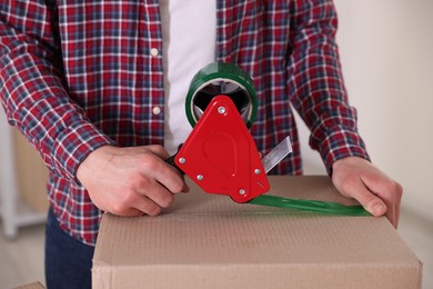 Photo of Man using dispenser with adhesive tape to seal cardboard box indoors, closeup