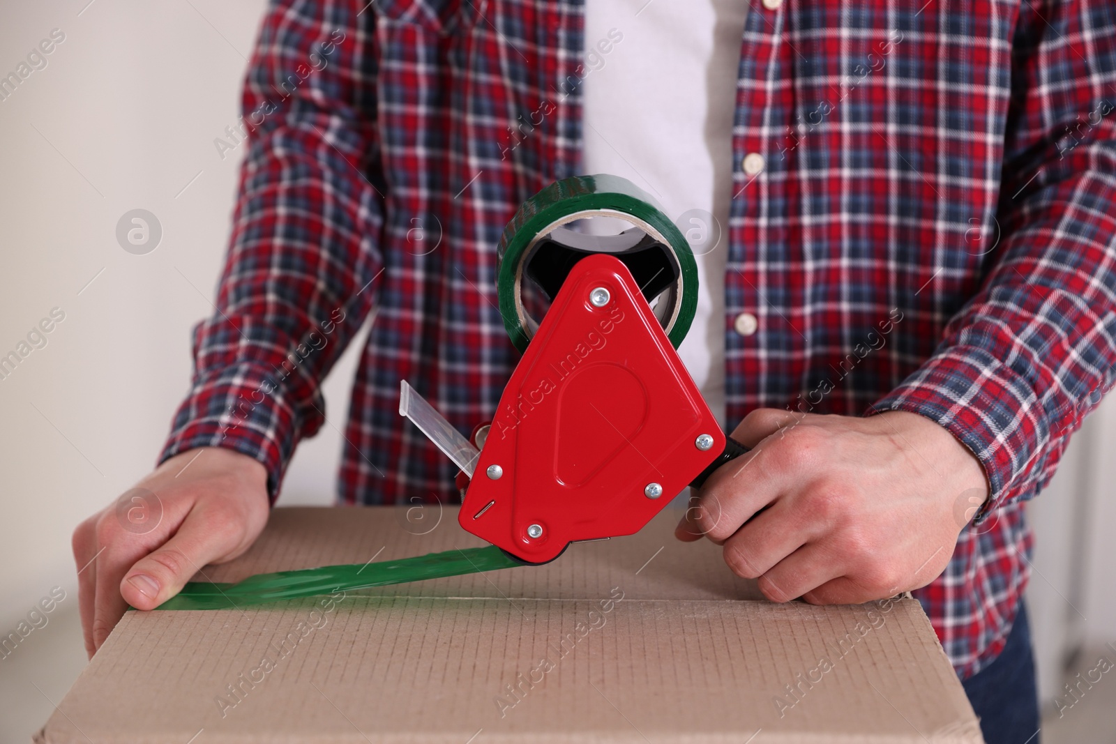 Man using dispenser with adhesive tape to seal cardboard box indoors, closeup Photo of Man using dispenser with adhesive tape to seal cardboard box indoors, closeup