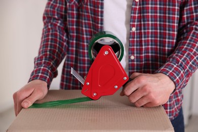 Photo of Man using dispenser with adhesive tape to seal cardboard box indoors, closeup