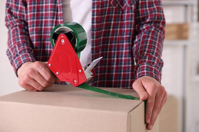 Photo of Man using dispenser with adhesive tape to seal cardboard box indoors, closeup
