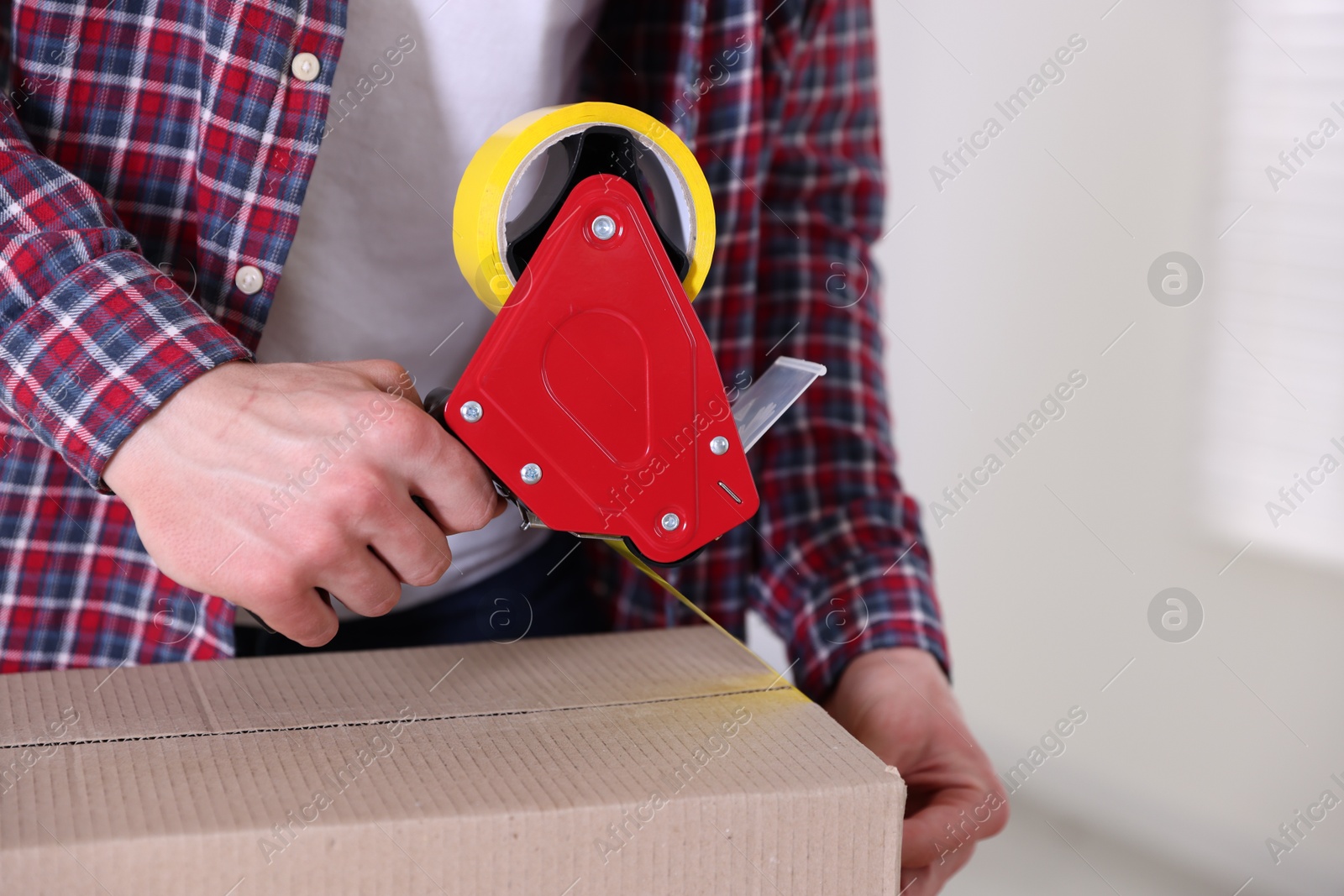 Man using dispenser with adhesive tape to seal cardboard box indoors, closeup Photo of Man using dispenser with adhesive tape to seal cardboard box indoors, closeup
